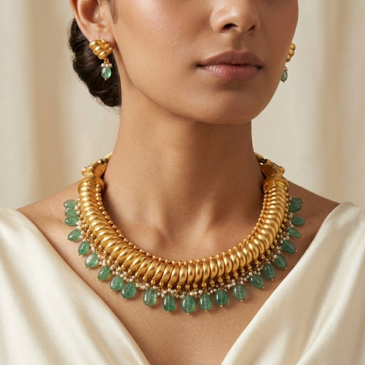 A close-up of a model wearing a gold-toned multi-strand choker necklace with green teardrop gemstones, matching drop earrings, and a cream satin top.