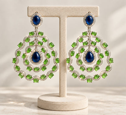 A pair of ornate drop earrings displayed on a cream jewelry stand. The earrings feature large, intricate circular filigree designs set with bright green marquise-cut stones and a deep blue teardrop center, surrounded by small clear pavé stones.