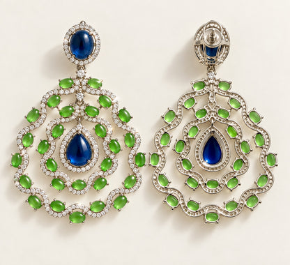 A pair of ornate silver-toned statement earrings featuring large oval and teardrop-shaped emerald-cut blue stones surrounded by rows of green marquise-cut stones and small clear accent stones, displayed on a light cream background.