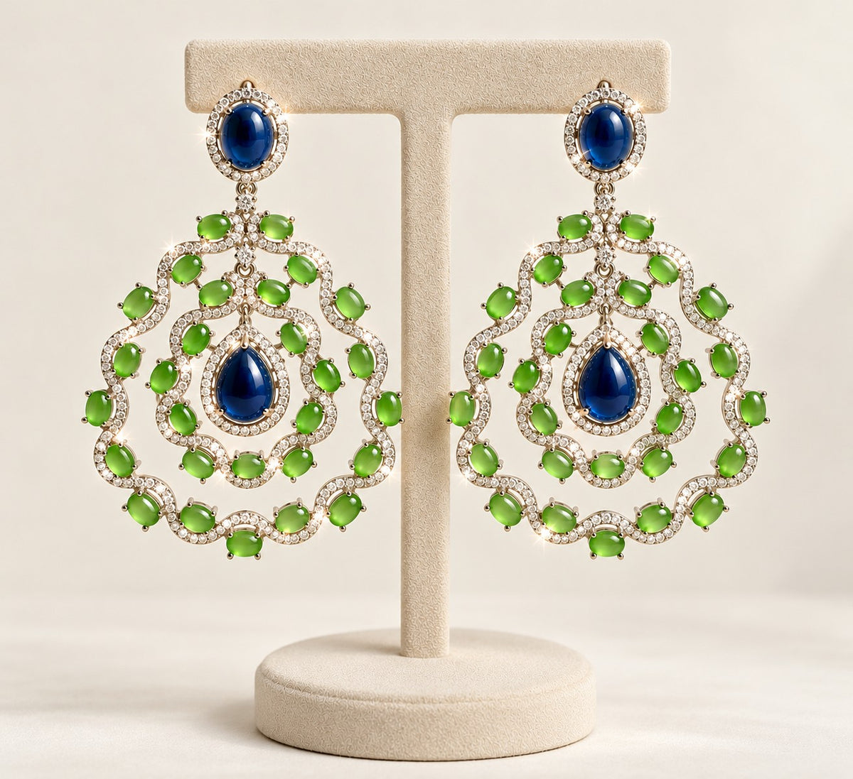 A pair of ornate drop earrings displayed on a cream jewelry stand against a soft beige background. Each earring features a large oval blue stone at the top and a scalloped circular design below with multiple green stones and small clear accent stones.
