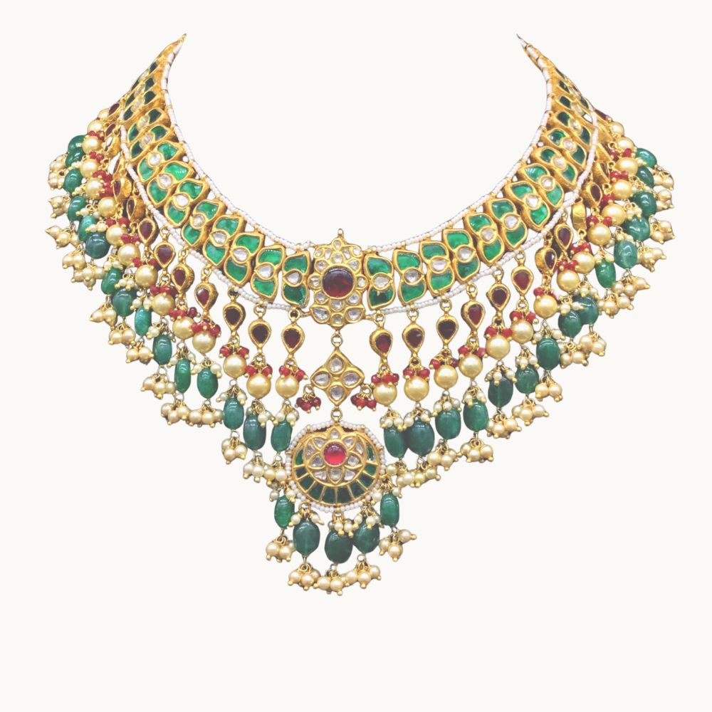 An ornate gold jadau traditional Indian  necklace decorated with green emerald-cut stones, red gemstone accents, and dangling gold bead chains, shown against a plain off-white background.