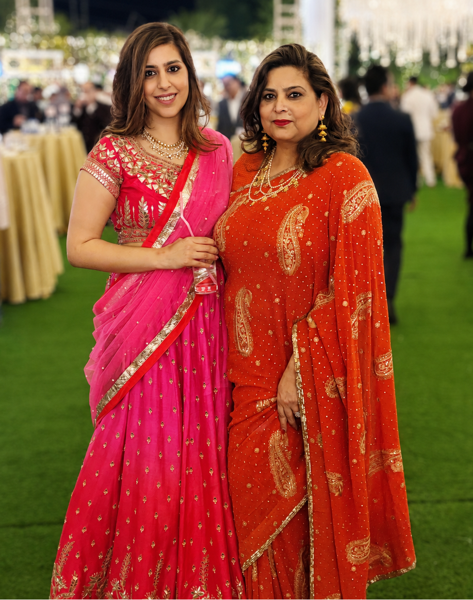 Two women posing in traditional Indian sarees at an outdoor evening event. The woman on the left wears a vibrant pink saree with gold embroidery, and the woman on the right wears a bright orange saree with gold paisley patterns.