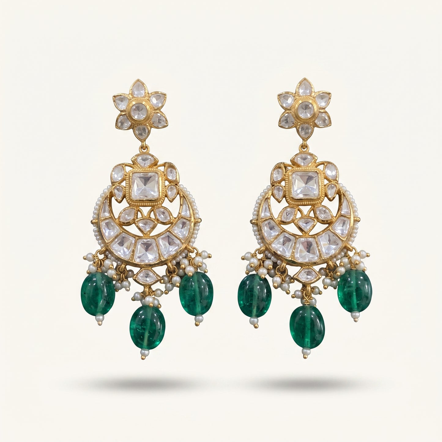 A pair of ornate gold-plated Indian Kundan chandelier earrings featuring clear kundan stones, small white pearl beads, and dangling emerald-cut green stones, shown against a light cream background.