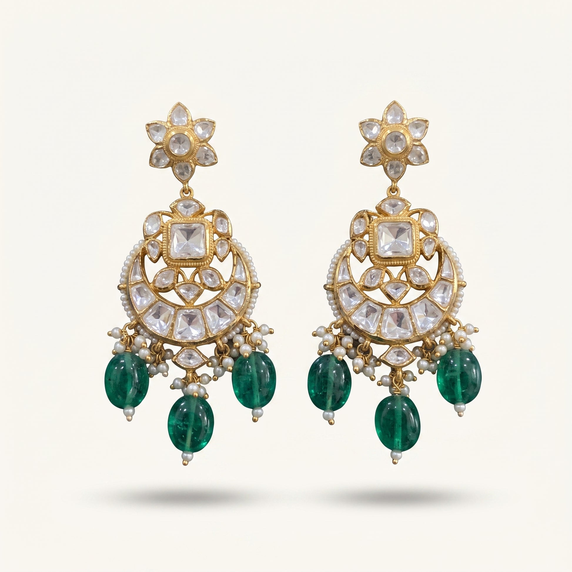 A pair of ornate gold-plated Indian Kundan chandelier earrings featuring clear kundan stones, small white pearl beads, and dangling emerald-cut green stones, shown against a light cream background.