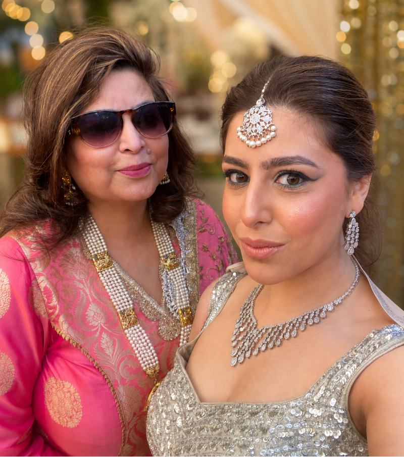 Two women posing closely together at what appears to be a festive event. The woman on the left wears a bright pink traditional outfit with gold embroidery, layered pearl necklaces, and sunglasses.