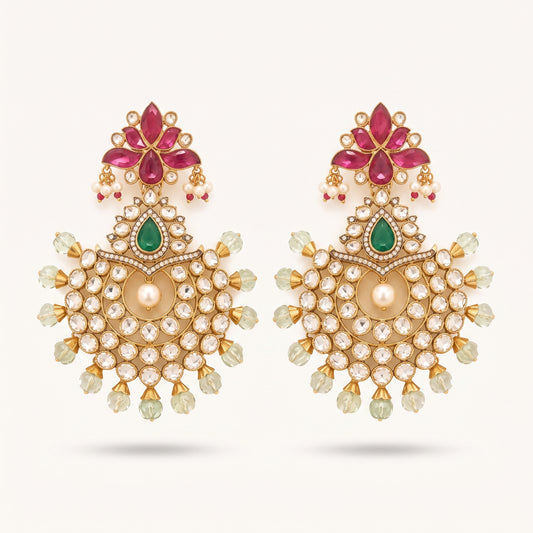 A pair of ornate gold-toned Indian chandelier earrings featuring pink gemstone floral motifs, green teardrop stones, and a central cream pearl surrounded by clear rhinestones and pale green crystal drops, set against a light cream background.