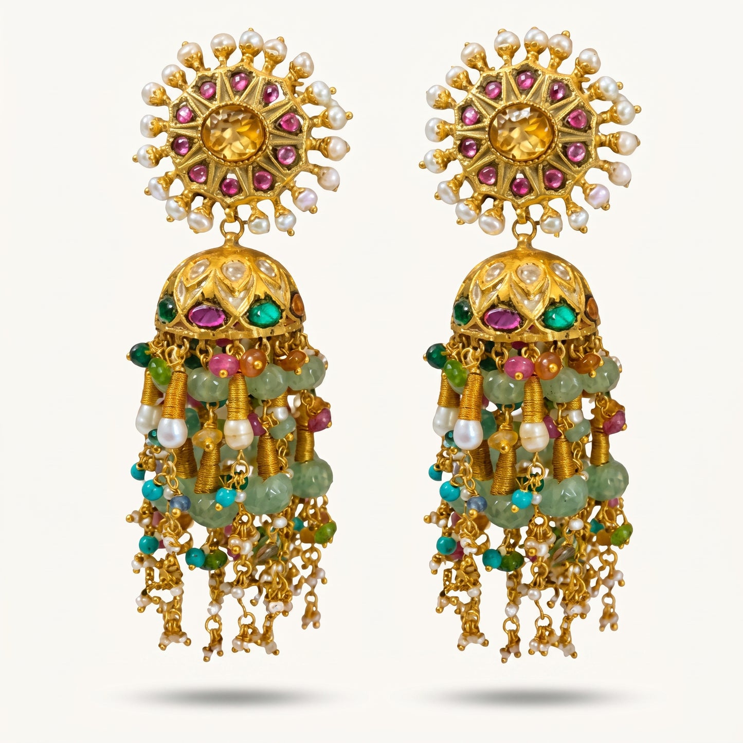 A pair of ornate gold-toned traditional Indian jhumka earrings embellished with white pearls, pink and green stones, and hanging green bead strands, set against a light off-white background.
