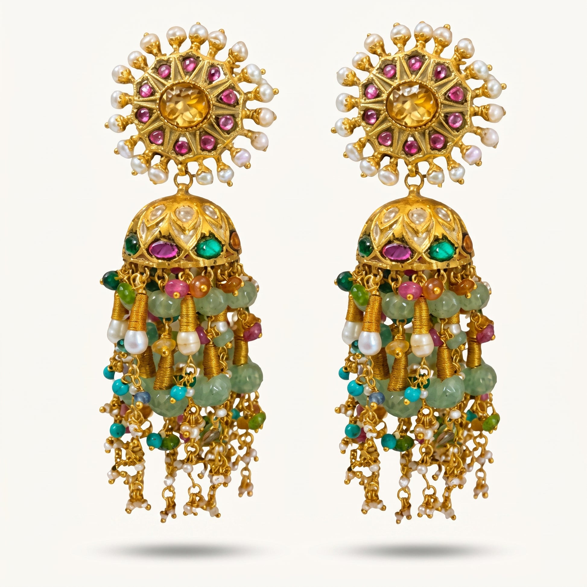 A pair of ornate gold-toned traditional Indian jhumka earrings embellished with white pearls, pink and green stones, and hanging green bead strands, set against a light off-white background.