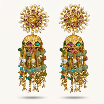 A pair of ornate gold-toned traditional Indian jhumka earrings embellished with white pearls, pink and green stones, and hanging green bead strands, set against a light off-white background.