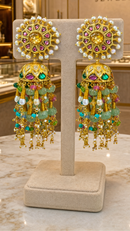 A pair of ornate gold-toned Indian jhumka earrings with multicolored gemstones and bead tassels, displayed on a beige jewelry stand inside a luxury jewelry store. The background features the brand name 'RATNALI JEWELS' on a beige wall.