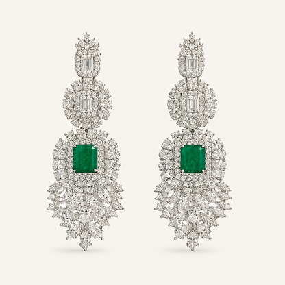 A pair of elaborate drop earrings featuring square emerald-cut green gemstones surrounded by dense clusters of clear baguette and round-cut stones in a silver-toned setting, presented on a light cream background.