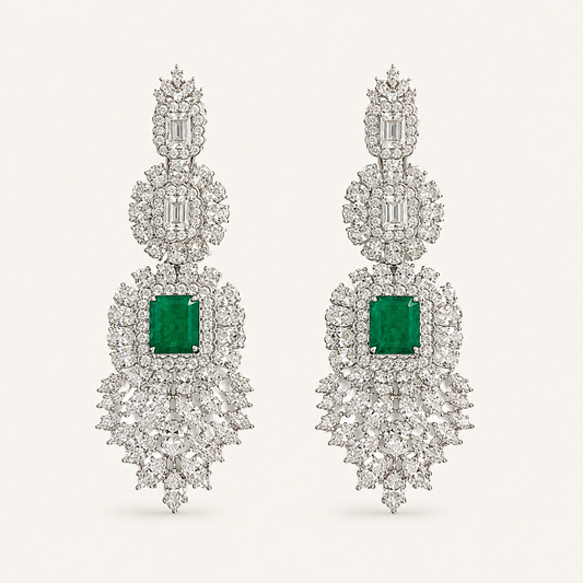 A pair of elaborate drop earrings featuring square emerald-cut green gemstones surrounded by dense clusters of clear baguette and round-cut stones in a silver-toned setting, presented on a light cream background.