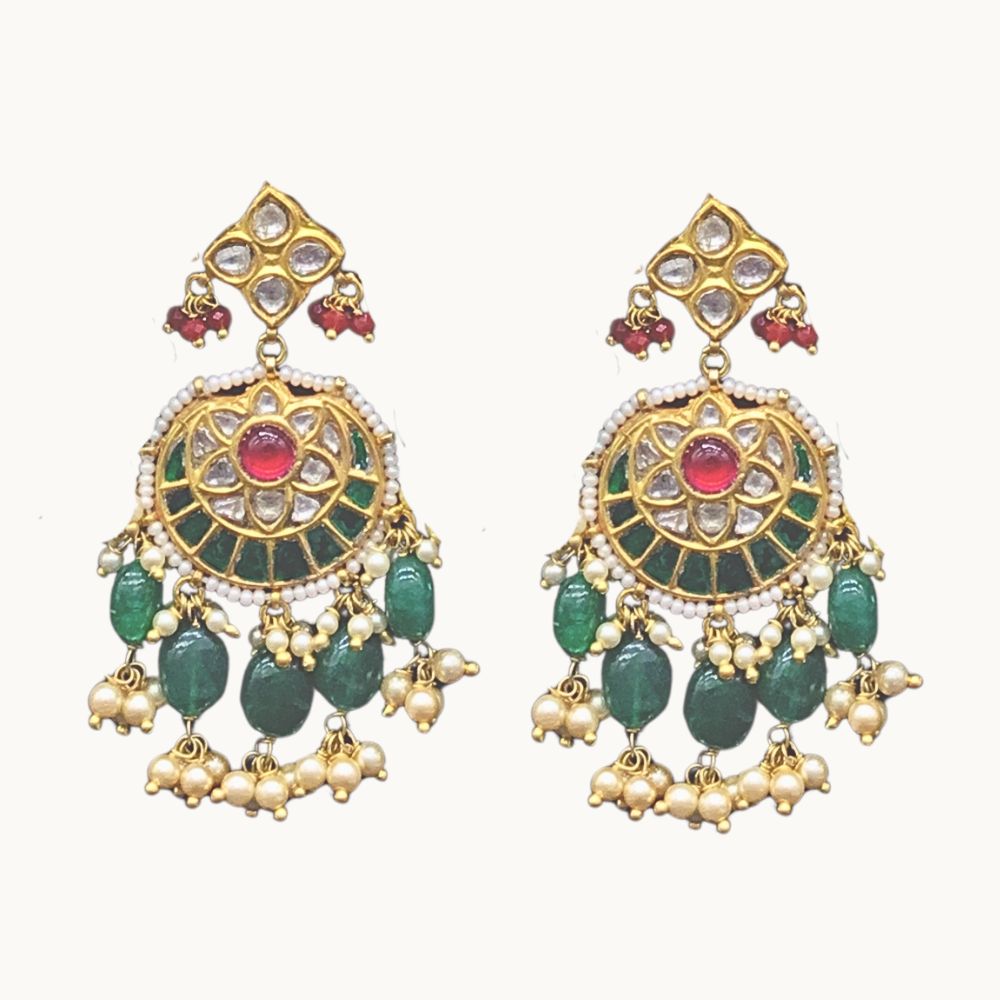 A pair of traditional Indian gold-plated jhumka earrings decorated with green stones, white faux pearls, and red gemstones, shown against a plain off-white background.