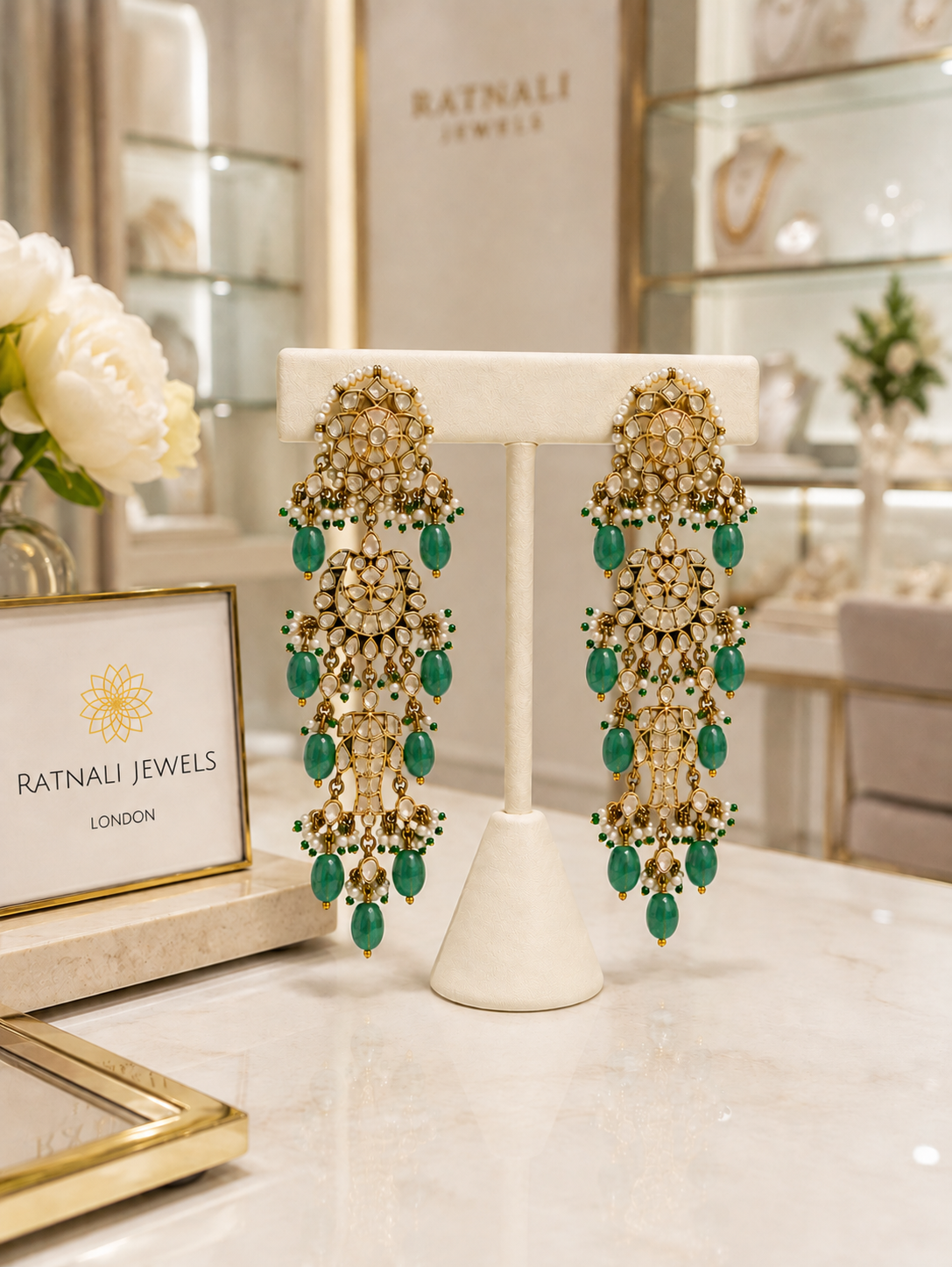 A pair of ornate gold and green drop earrings displayed on a white jewelry stand on a marble counter inside a Ratnali Jewels boutique.