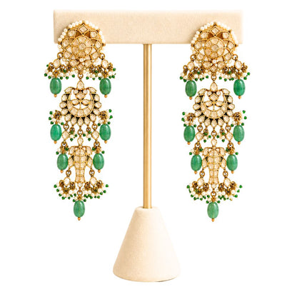 A pair of ornate gold-toned chandelier earrings with green bead accents, displayed on a cream-colored jewelry stand against a white background.