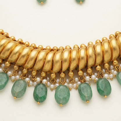 A close-up of a gold-toned statement necklace featuring a thick textured collar, dangling gold beads, small white pearls, and faceted green gemstone drops on a light cream background.