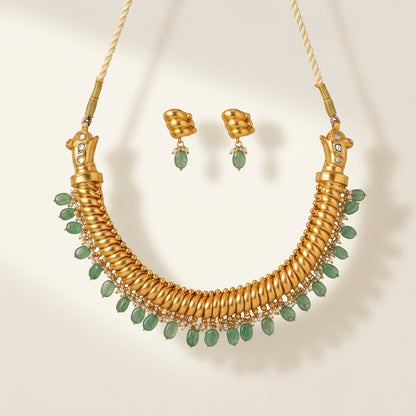 Gold-toned statement necklace and matching drop earrings with green teardrop gemstones and small clear stones, displayed on a warm cream background.