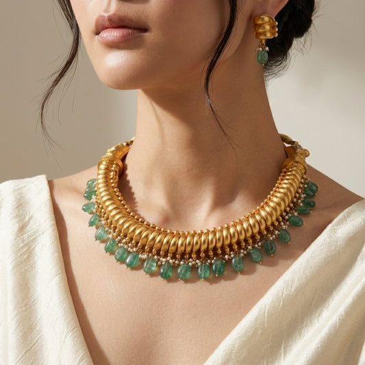 A close-up of a model wearing a chunky gold collar necklace with dangling green teardrop gemstones, matching gold drop earrings, and a cream-colored top.