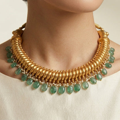 A close-up of a model wearing a chunky gold collar necklace adorned with dangling green teardrop gemstones and small gold beads, paired with a white top.