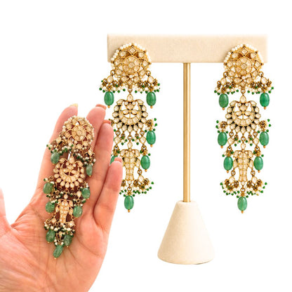 A pair of ornate gold-toned dangle earrings with green bead accents, displayed on a cream jewelry stand, with a hand holding a matching single earring for scale against a white background.